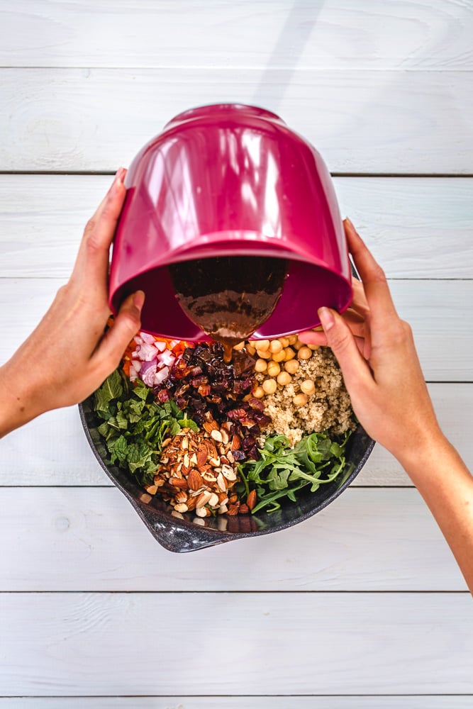 Hands pour ingredients from a pink bowl into a skillet filled with quinoa, chickpeas, arugula, nuts, dried fruit, onion, and herbs on a white wooden surface.