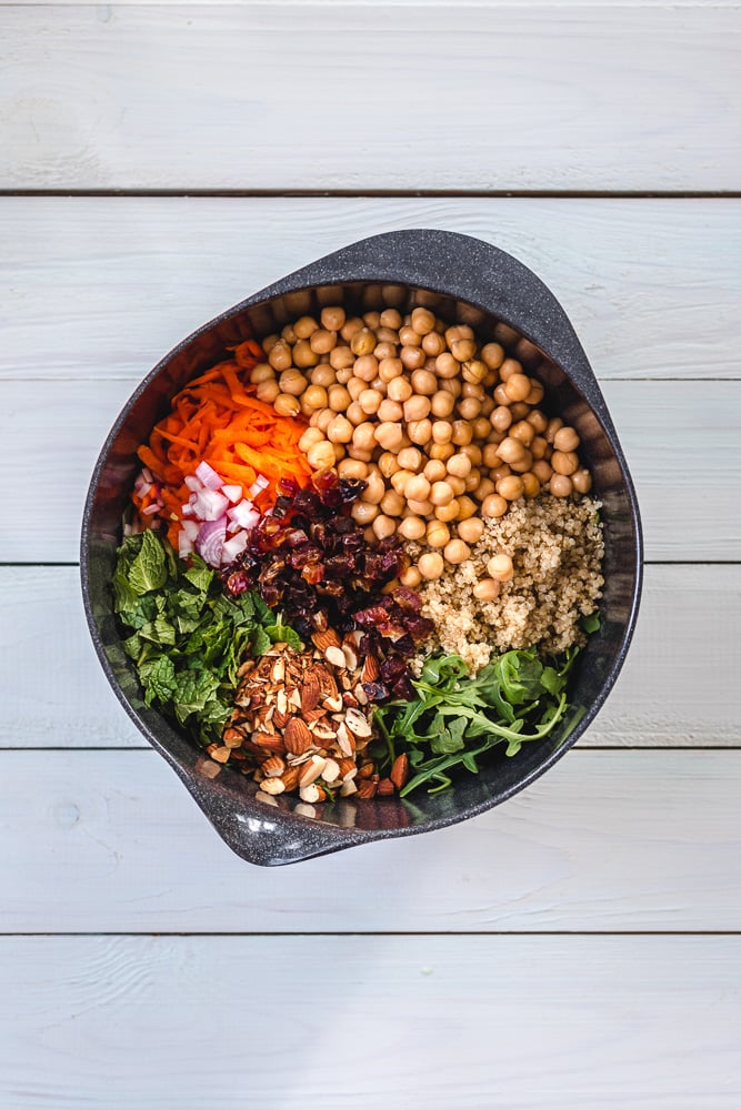 A bowl with chickpeas, quinoa, shredded carrot, chopped red onion, dried cranberries, almonds, mint, and arugula on a light wooden table.