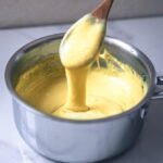 Image of melty and stretchy vegan mozzarella in a saucepan with a wooden spoon scooping out the vegan cheese to show its cheesy consistency.