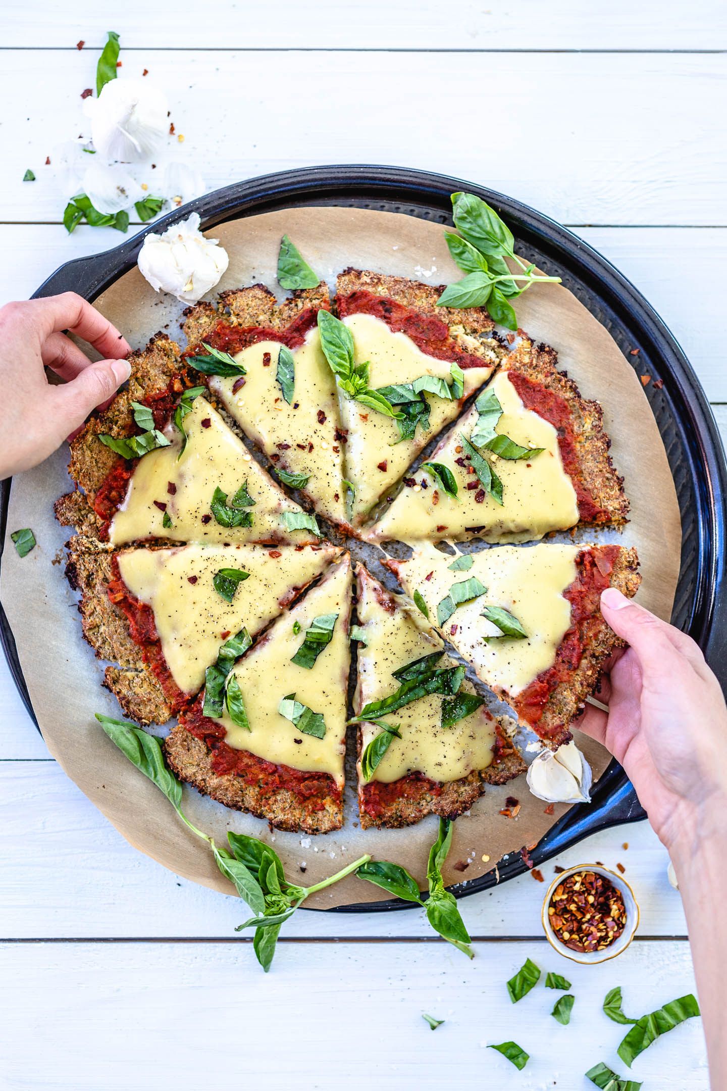 Cauliflower pizza crust with marinara, homemade vegan mozzarella and fresh basil with hands picking out slices. The pizza is on parchment paper with sprinkles of basil sitting on a black plate. The parchment paper is sitting on a white backdrop, a cup of chili flakes  in the bottom right and a head of garlic on opposite sides of the pizza. 