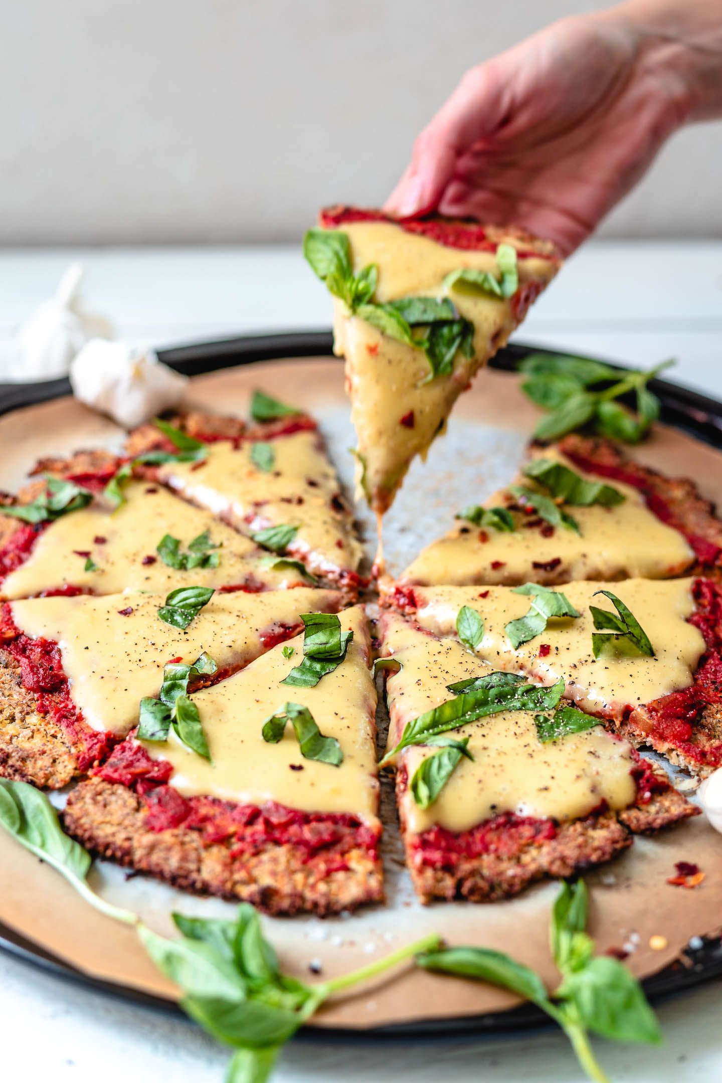 Cauliflower pizza crust with marinara, homemade vegan mozzarella and fresh basil, a hand is picking up a slice of the pizza to show the texture. The pizza is on parchment paper with sprinkles of basil. The parchment paper is sitting on a white backdrop and a head of garlic in the top left. 