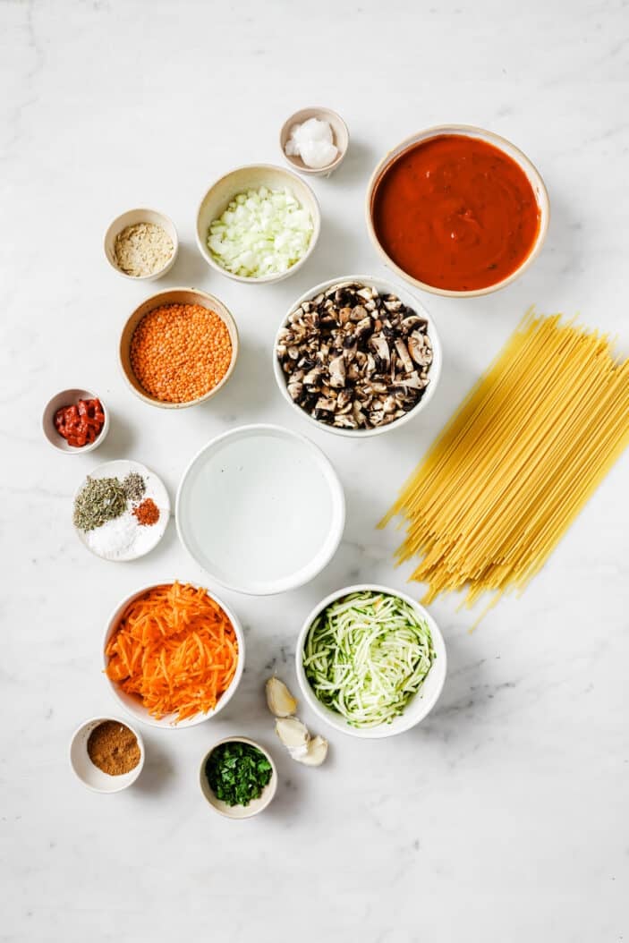 all of the ingredients required to make lentil bolognese sauce separated into individual bowls with dry spaghetti noodles on the side
