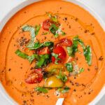 close up of tomato gazpacho that has been trasnfered to a white bowl and decorated with fresh tomatoes, chopped basil, freshly ground pepper and olive oil to drizzle