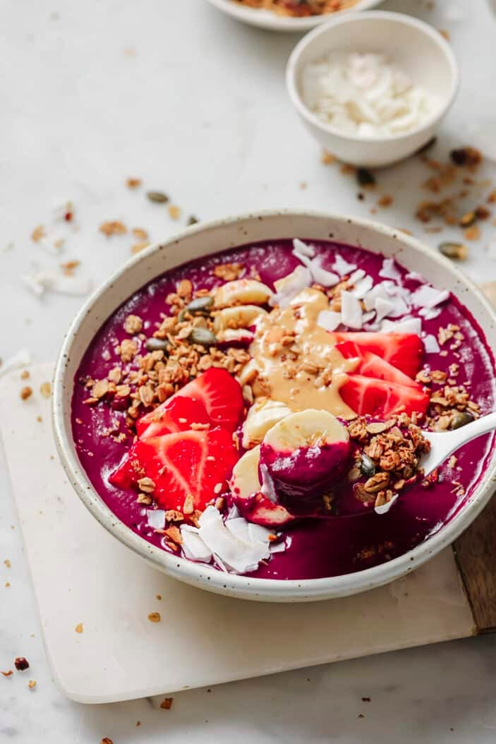 thick and creamy acai bowl topped with fresh strawberries, bananas, granola and coconut flakes. Sitting on a wood cutting board on a white backdrop.