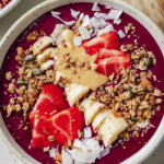 close up of thick and creamy acai smoothie in a white bowl topped fresh strawberries, bananas, granola and coconut flakes. Sitting on a wood cutting board on a white backdrop.