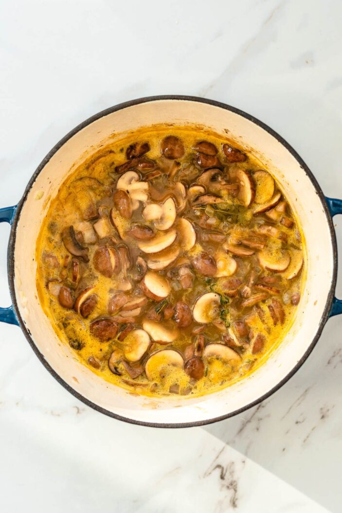 Simmering cream of mushroom soup in a large pot on a marble cutting board. The sliced cremini mushrooms are in a brothy mixture in the pot.