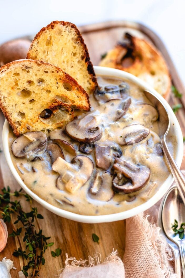A white bowl filled with creamy and chunky vegan mushroom soup with two slices of toast in the top corner of the soup. The bowl of soup is on a wood cutting board. There is fresh thyme on the board and a kitchen towel.