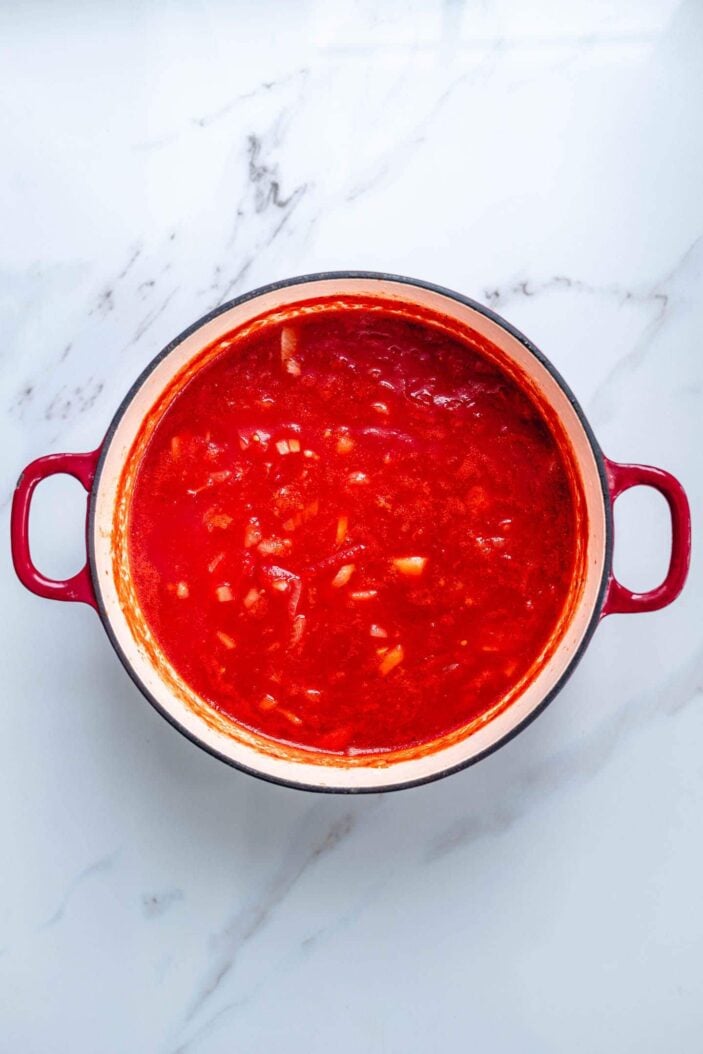 Onions, garlic, vegetable broth and canned tomatoes. This mixture is in a red dutch oven. The dutch oven is sitting on a white backdrop.