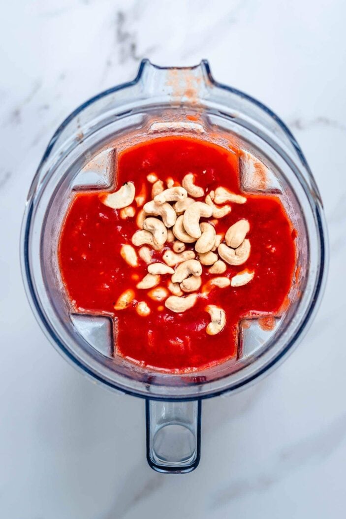 The tomato mixture and raw cashews added to a blender container. The container is sitting on a white backdrop.