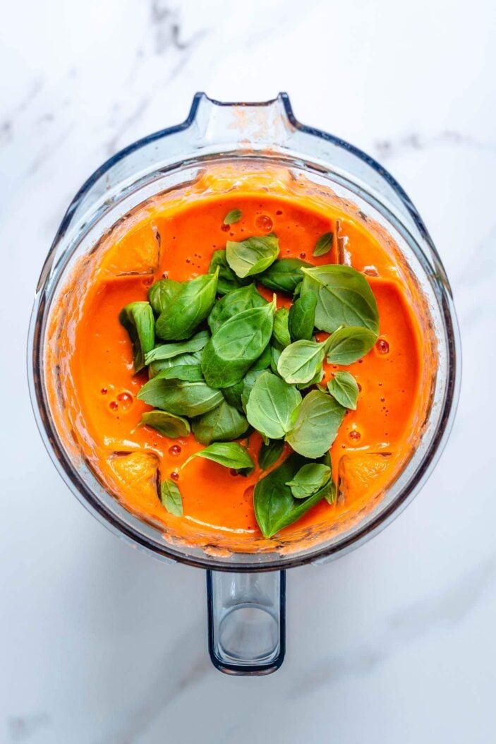 The tomato soup mixture in a blender container with basil. The blender container is sitting on a white backdrop.