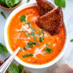 Vegan tomato basil soup topped with sliced basil and two slices of toast. The soup is poured into a white bowl with a silver spoon inside. The bowl is sitting on a white backdrop with clumps of basil and a towel in the bottom right.