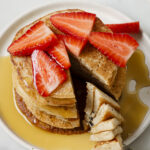 Fluffy coconut flour pancakes topped with strawberries, maple syrup and cut to show the fluffy interior. The pancakes are stacked on to a white plate. The plate is sitting on a white backdrop.