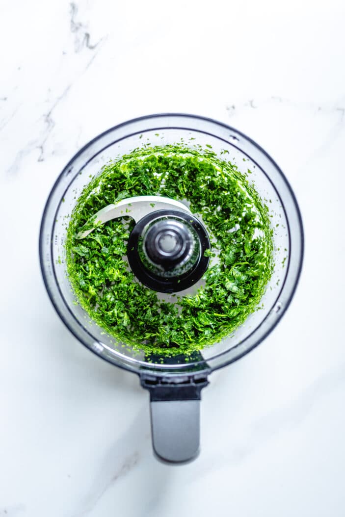 fresh herbs pulsed together until finely chopped in a food processor. The food processor is on a white backdrop.
