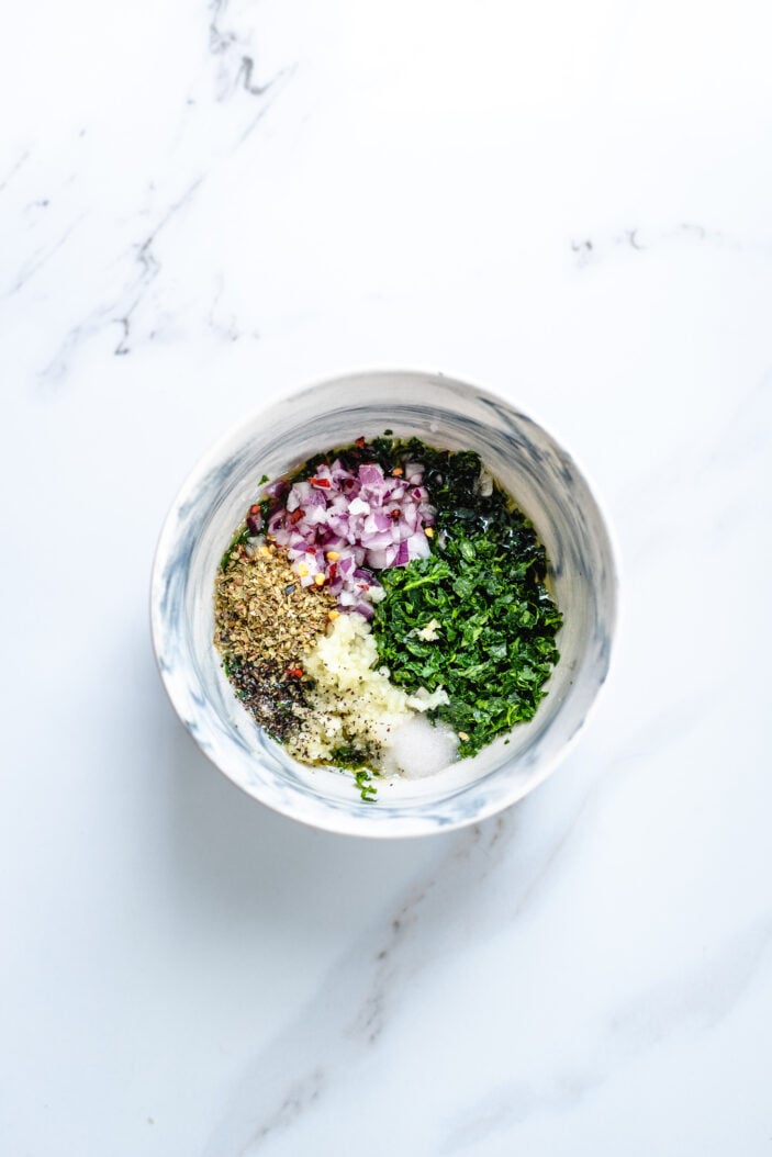 Chopped onion, garlic, dreid herbs and salt added to a bowl of fresh herbs on a white backdrop.