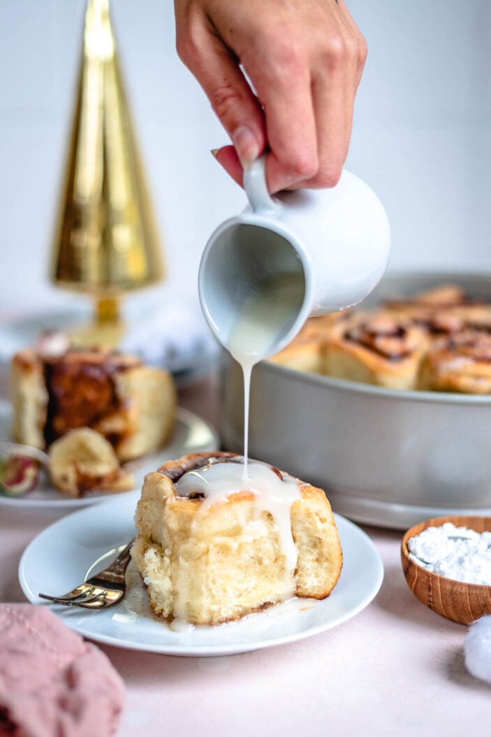 vegan cinnamon rolls being drizzled with homemade icing. There are cinnamon rolls in the background and the white plate is on a pink backdrop.