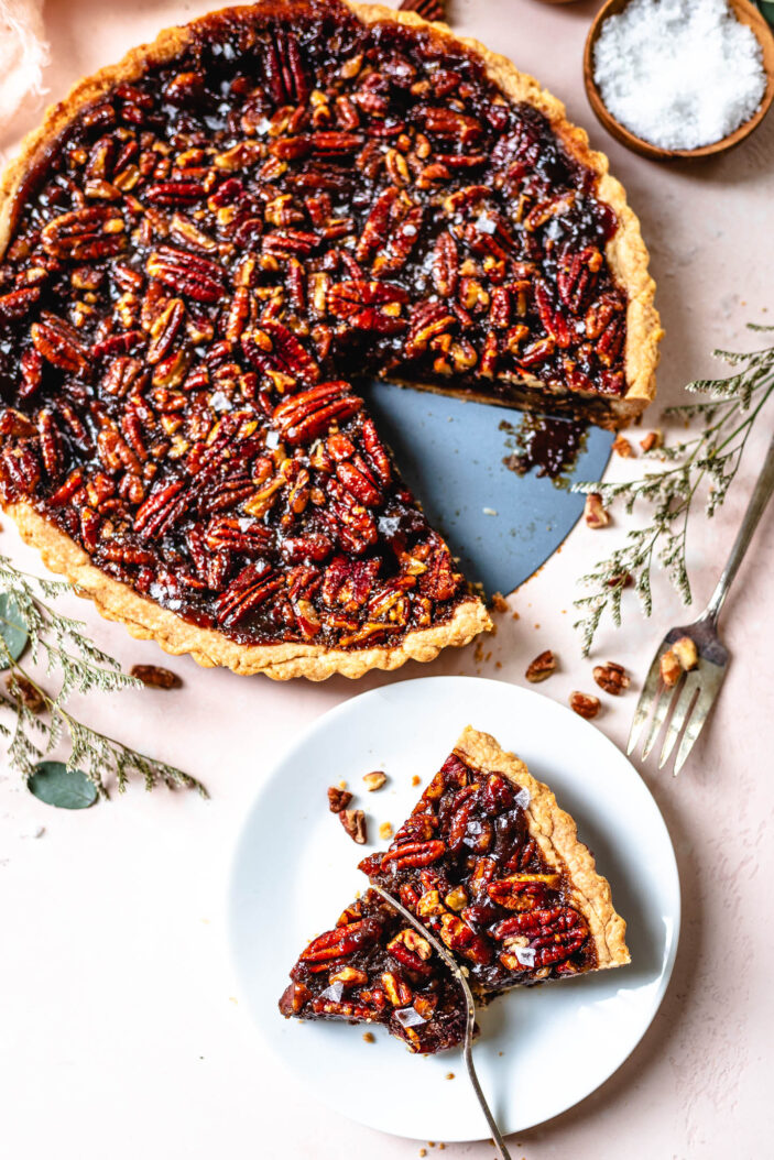 A pecan pie with a slice removed sits on a table, with the slice placed on a white plate nearby. A fork rests on the plate, and some pecans are scattered around.