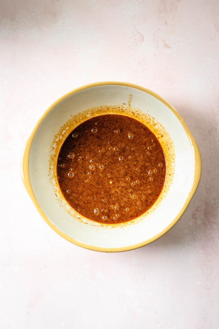 A white bowl with a tan rim holds a brown, bubbly liquid mixture, viewed from above on a light pink background.