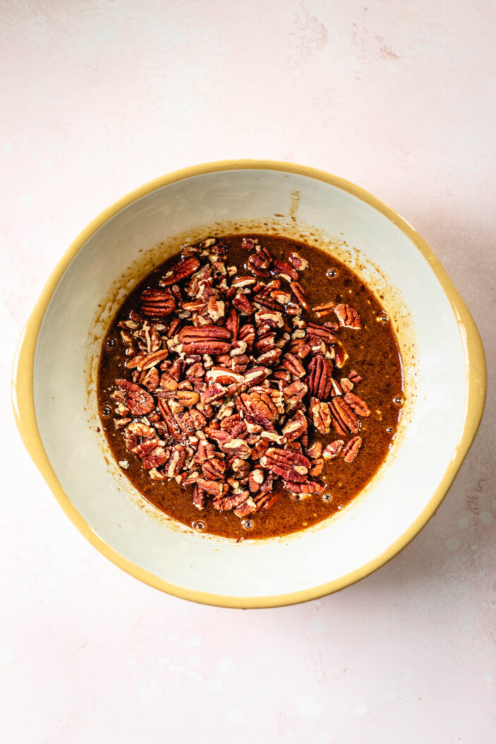A ceramic bowl filled with chopped pecans and a brown spiced liquid mixture, placed on a light pink surface.