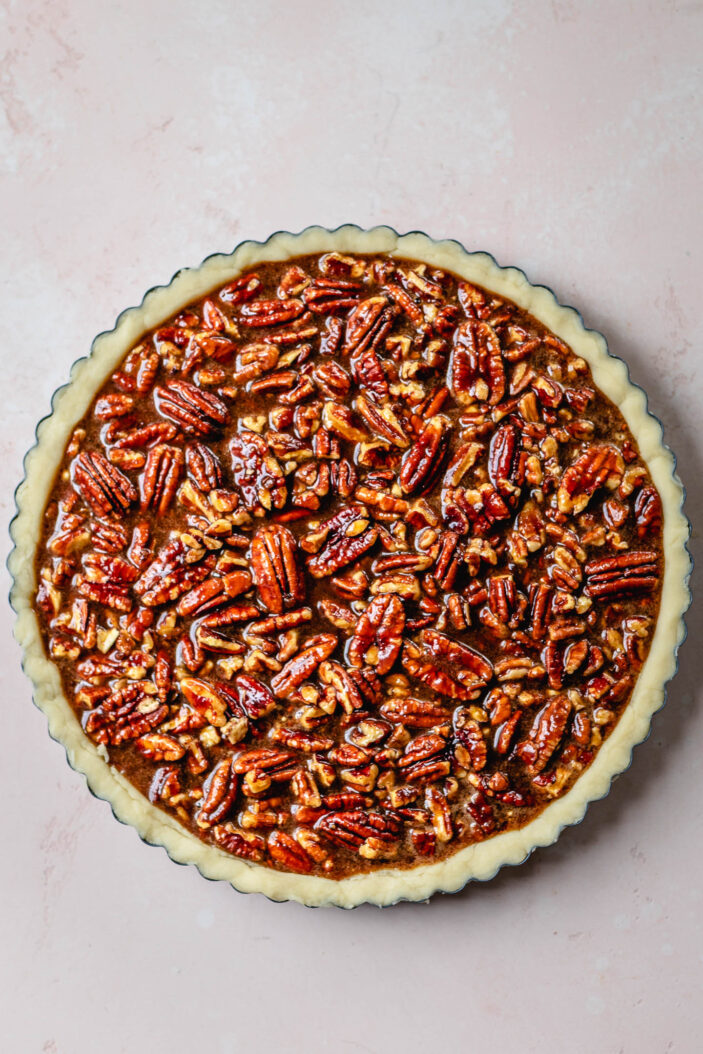 A pecan pie in a round baking pan, filled with glossy whole pecans on top of a golden crust, photographed from above against a light pink background.