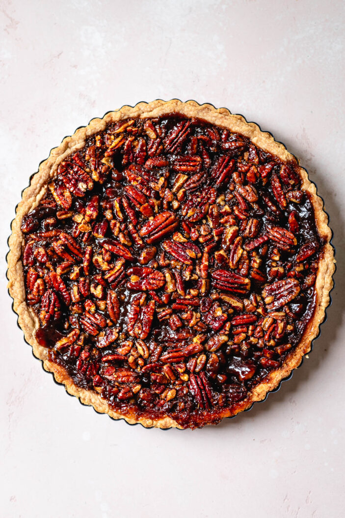 A whole pecan pie with a golden crust, filled with glossy, caramelized pecans, sits on a light pink surface.