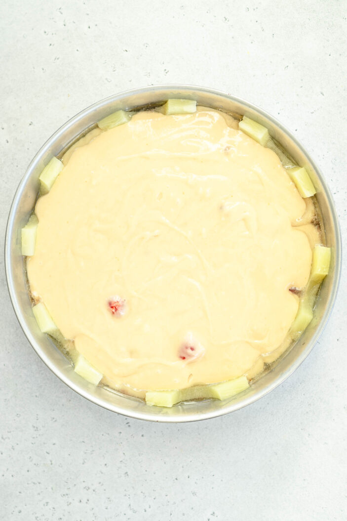 A round baking pan filled with cake batter, with pineapple chunks arranged along the edge of the pan on a light gray surface.