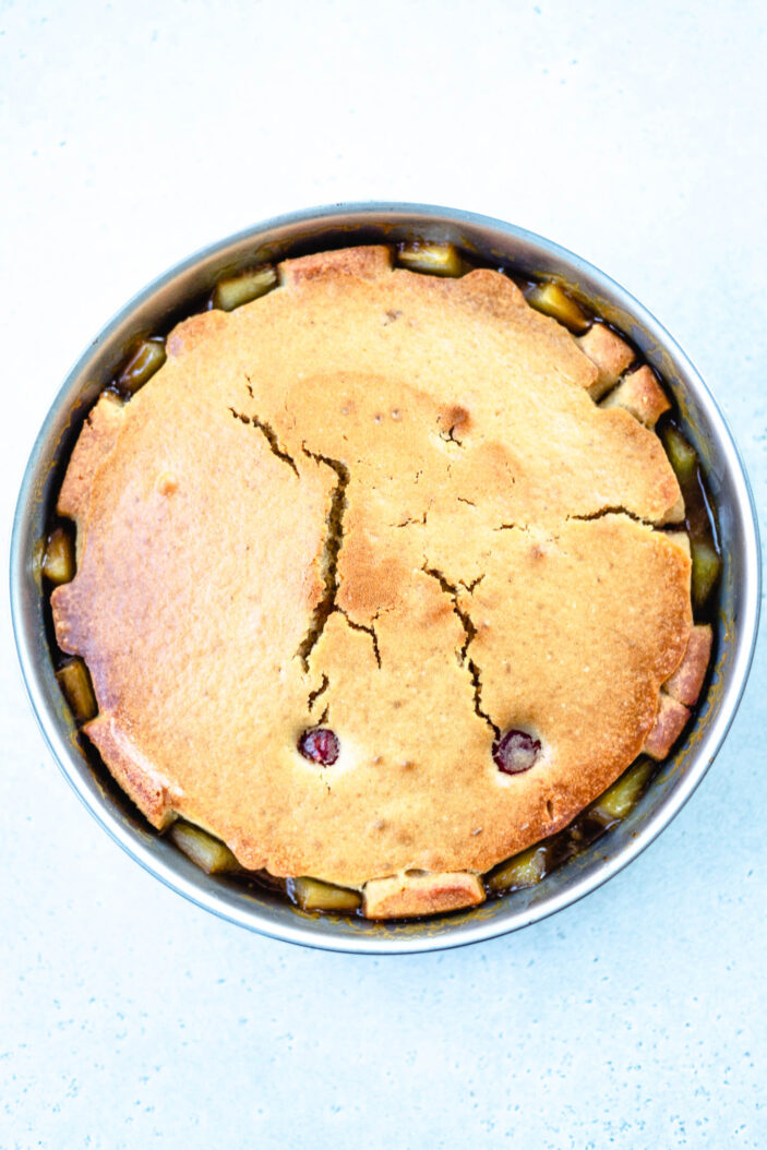 A round fruit cobbler with a golden-brown, cracked crust and pieces of fruit visible around the edges, baked in a metal pan on a light surface.