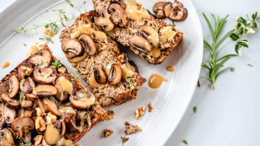 nut roast recipe with two slices laying down on a white oval plate, topped with mushrooms and vegan gravy