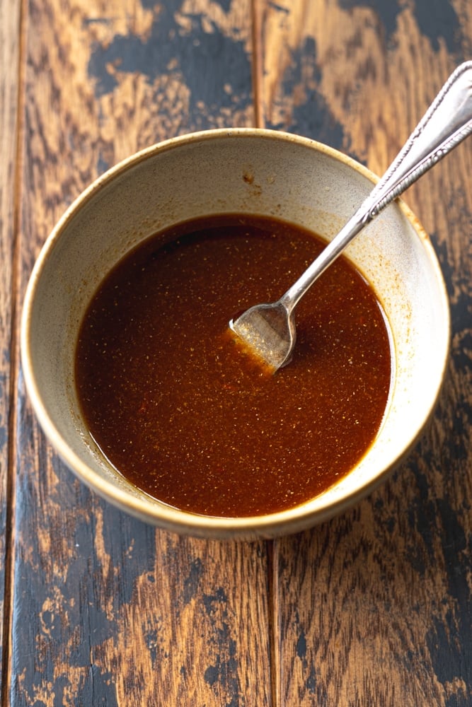 A bowl of dark reddish-brown sauce with a spoon resting inside, placed on a rustic wooden surface.