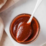 homemade vegan bbq sauce in a glass jar with a silver spoon spooning out some of the bbq sauce.