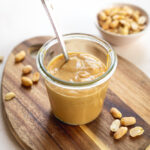 Peanut sauce in a clear glass jar with a silver spoon. Sitting on a wood cutting board.