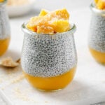 mango coconut chia pudding with chunks of coconut on top, sprinkled with toasted shredded coconut. The jars are sitting on a white cutting board with a white backdrop.