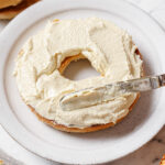 image of tofu cream cheese being spread onto a bagel on a white plate. The plate is sitting on a white backdrop.