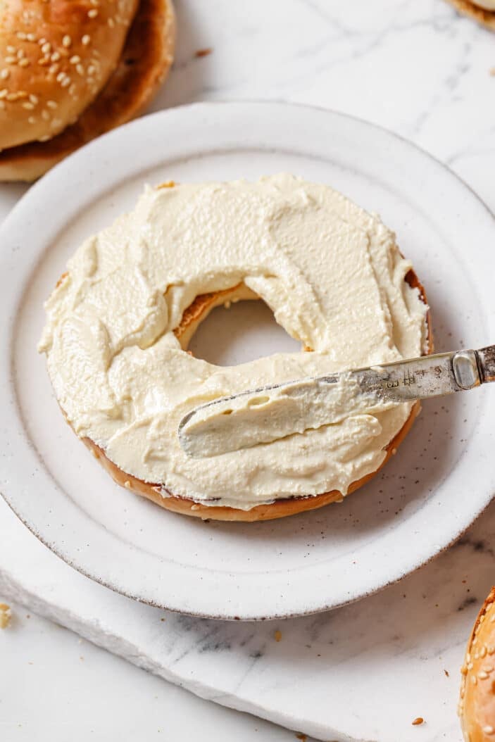 image of tofu cream cheese being spread onto a bagel on a white plate. The plate is sitting on a white backdrop.