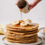 almond flour pancakes in a stack on a white plate with a white backdrop. Almond flour pancakes are topped with sliced banana and are being drizzled with maple syrup.