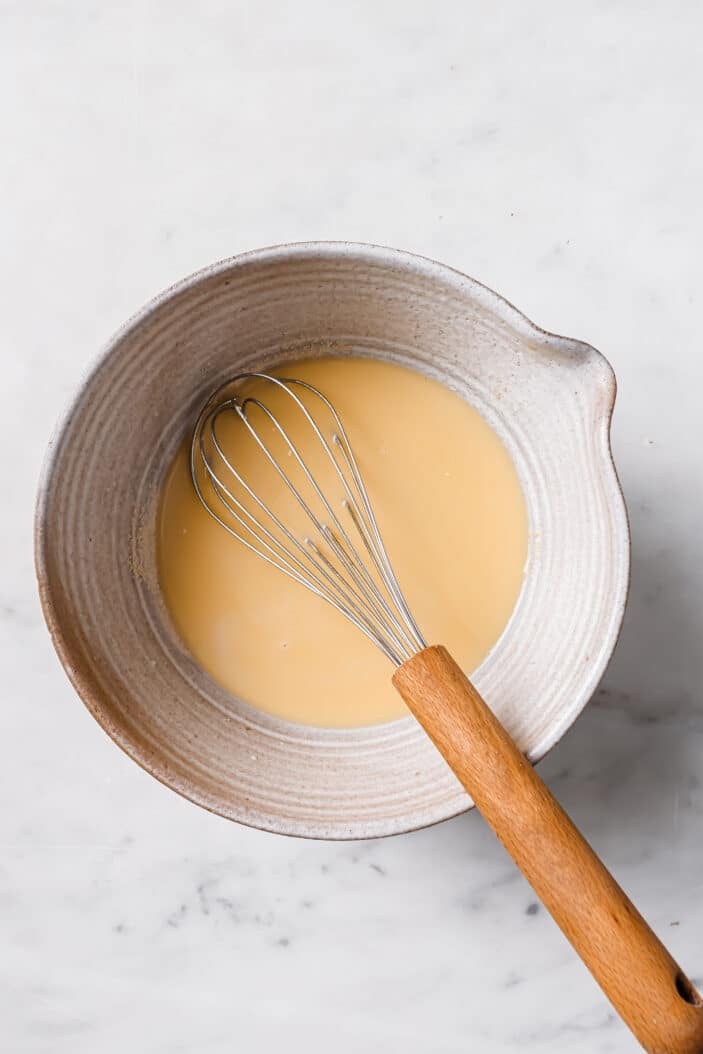 Heated almond milk, vegan butter and sugar poured into a medium grey bowl with a whisk in the middle. The bowl is sitting on a white backdrop.