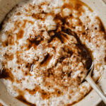 Cinnamon Roll Oatmeal topped with cinnamon butter. The oatmeal is in a white bowl with a spoon in the middle. The bowl is sitting on a wooden backdrop.