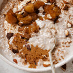 Apple pie oatmeal topped with coconut yogurt, almond butter, almonds and cinnamon. The oatmeal is in a white bowl with a spoon in the oatmeal to show how creamy it is. The bowl is sitting on a white backdrop with sprinkles of almonds and cinnamon.