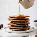 Stacked buckwheat pancakes topped with a dollop of butter on a white place sitting on top of a white cutting board with white countertop and white backdrop. There is a small jug of maple syrup overtop of the pancakes that's being drizzled onto the top of the pancakes.