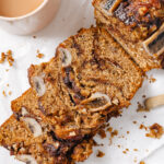 Cinnamon Swirl Banana Bread, topped with pecans and caramelized banana sliced to show the moist interior. The banana bread is sitting on parchment paper. The parchment paper is laid out on a white backdrop with a cup of coffee in the top left.