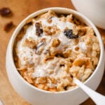 carrot cake oatmeal in a white bowl topped with coconut yogurt, raisins and walnuts. A silver spoon is in the bowl. The bowl is sitting on a round wood cutting board on a white backdrop. There's raisins and walnuts sprinkled beside the bowl on the cutting board.