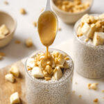peanut butter being drizzled on top of the chia pudding with a silver spoon. The chia pudding is in a glass and is also topped with chopped banana and chopped peanuts. The two chia puddings in glasses are sitting on a wood and marble cutting board on a white backdrop.