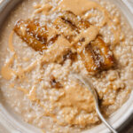 Vegan millet porridge topped with peanut butter, caramelized bananas and coconut flakes. The porridge is in a white bowl that is placed on a grey plate with a silver spoon in the middle. The bowl is sitting on a white backdrop.