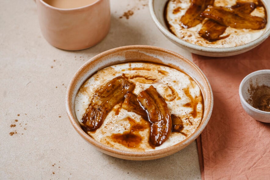 Image of a bowl of coconut yogurt with caramelized bananas on top. The bowl is sitting on a grey backdrop and there is another bowl of coconut yogurt with caramelized bananas in the back right hand corner. There is also a cup of tea in the top right hand corner. 