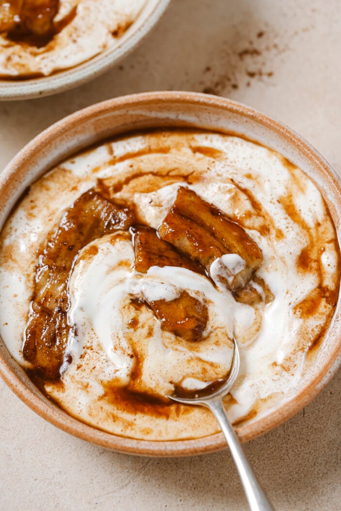 Close up image of a bowl of coconut yogurt with caramelized bananas on top. There is a silver spoon digging into the bowl. The bowl is sitting on a grey backdrop.