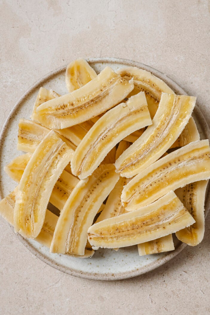 Image of bananas on a plate. The bananas have been halved and sliced and are sitting on the plate cut side up. The plate is sitting on a grey backdrop.