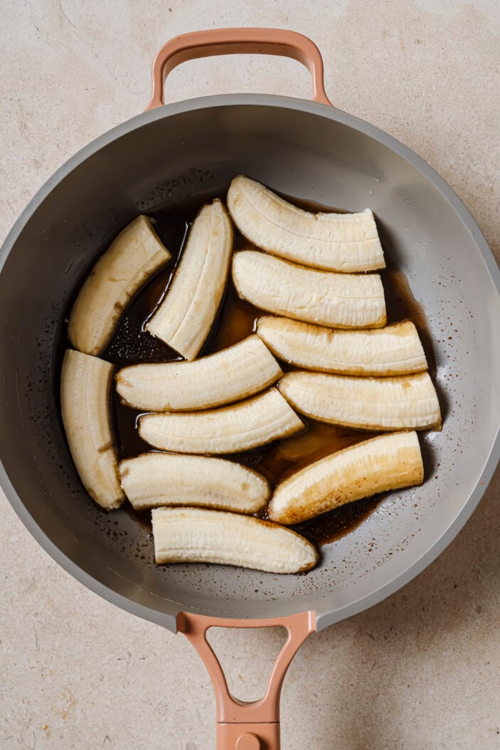 Image of the cut bananas in the skillet cut side down with the melted coconut oil and coconut sugar. The skillet is sitting on a grey backdrop.