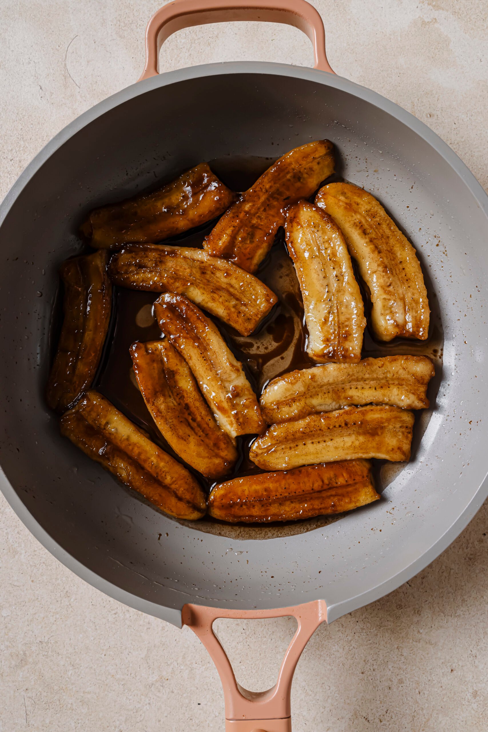 Image of the caramelized bananas in a skillet that are cut side up. The bananas are golden brown from being pan-fried. The skillet is on a grey backdrop. 