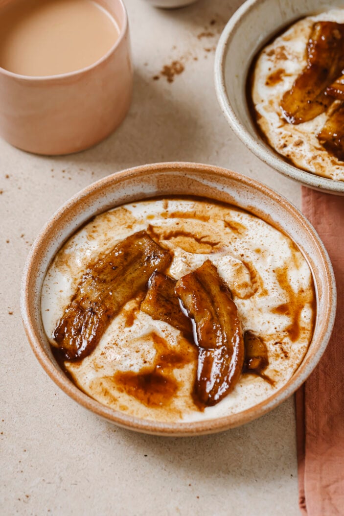 Image of caramelized bananas and coconut yogurt in a brown bowl on a light brown backdrop. There is a cup of coffee in the background as well as another bowl of caramelized bananas and coconut yogurt in the top right hand corner.