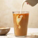 A glass cup of iced coffee with oat creamer being poured into the coffee. The glass cup is on a wooden cutting board with a creamy backdrop. There is a bowl of oats beside the glass of iced coffee.