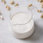image of vegan cashew cream in a clear pouring class with cashews sprinkled over a white countertop.